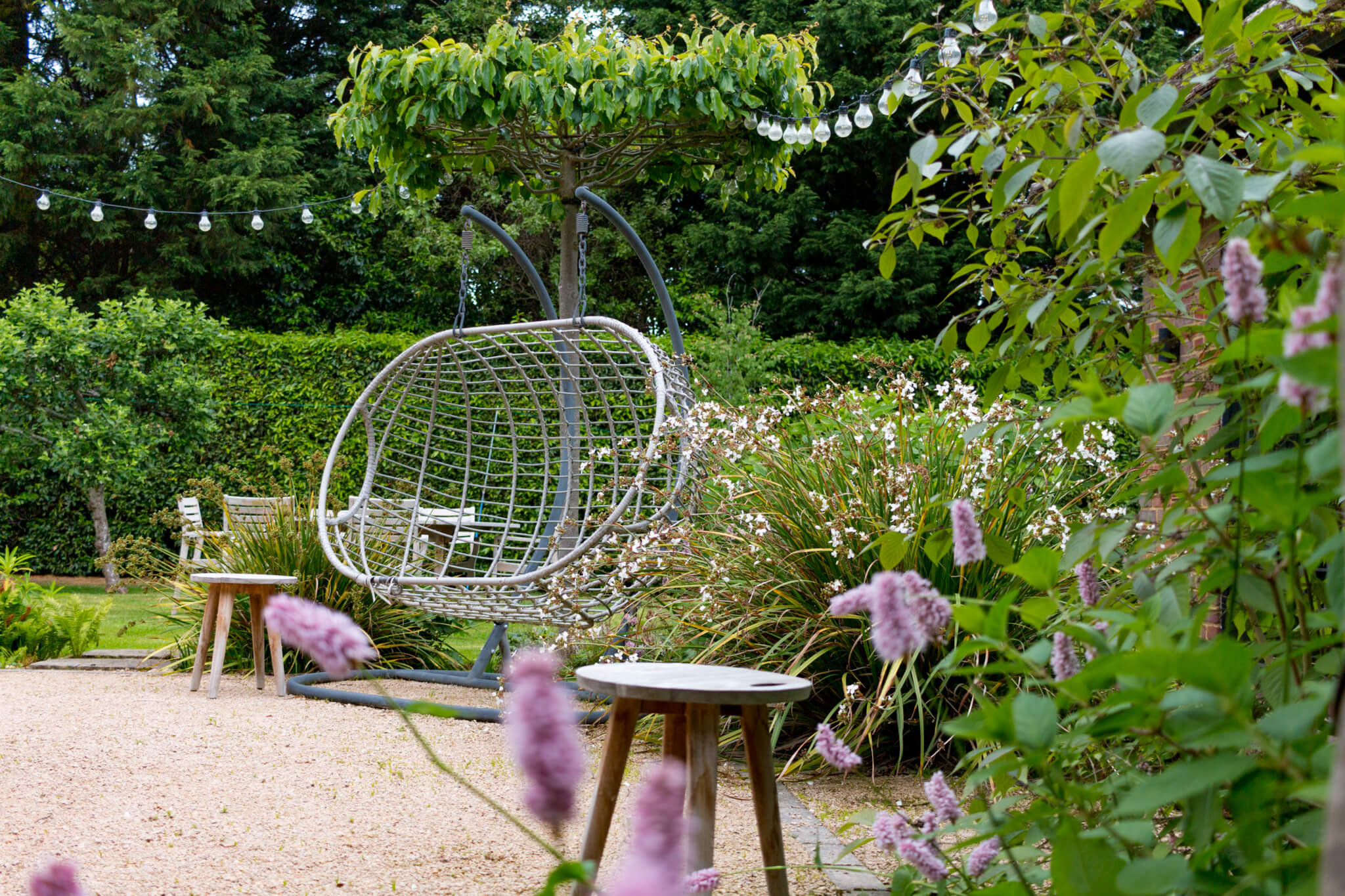 A metal swinging chair on a gravel surface, backdropped by some plants and a large hedge.